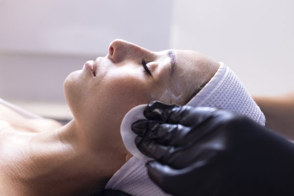 Close up of beautiful young brunette woman during facial cleansing and massage. Cosmetic procedures in a beauty salon or clinic. Lighted candles on background. Skin care and health concept.