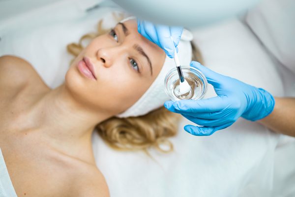 Hands of female cosmetologist applying a transparent mask with a brush on a woman face. Face skin care